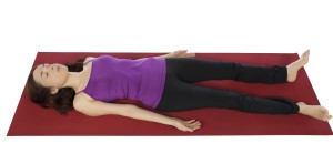 A person lying on a red yoga mat in corpse pose, wearing a purple top and black pants, embodies self-care practices for chronic pain relief, with arms gently resting by their sides.