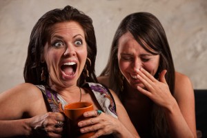 Two women seated together, one holding an orange mug and looking surprised, the other covering her mouth and laughing, perhaps sharing a moment of relief through CRPS coping techniques.