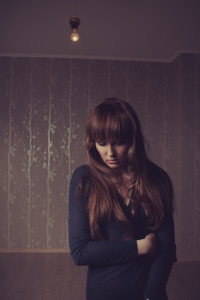 In a dimly lit room with patterned wallpaper and a bare light bulb above, a person with long hair in a dark sweater reflects on self-help resources for chronic illness, seeking understanding amid the shadows.