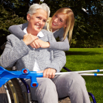 A woman in a wheelchair with crutches on her lap smiles as another woman stands behind her, embracing her. They are outdoors in a park setting, exemplifying self-care practices for chronic pain and showcasing warmth and support.