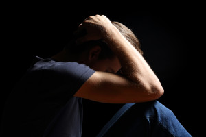 A person sits with their head in their hands, resting on their knees against a dark background, contemplating CRPS coping techniques to find relief and strength.