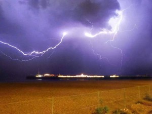 A dramatic lightning storm lights up the night sky over a ship in the distance, mirroring the electrifying resilience found in CRPS coping techniques, with a sandy foreground and a sturdy fence symbolizing self-help resources for chronic illness.